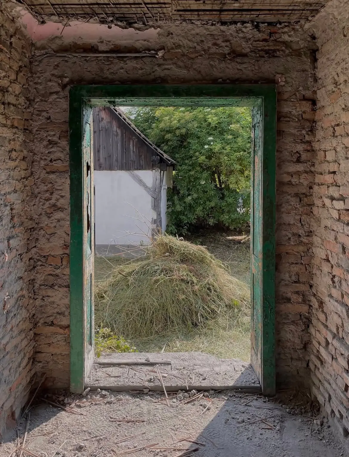 The northern entrance of the house seen from inside: a green-painted timber door frame, brick walls stripped of render, hay in the yard.
