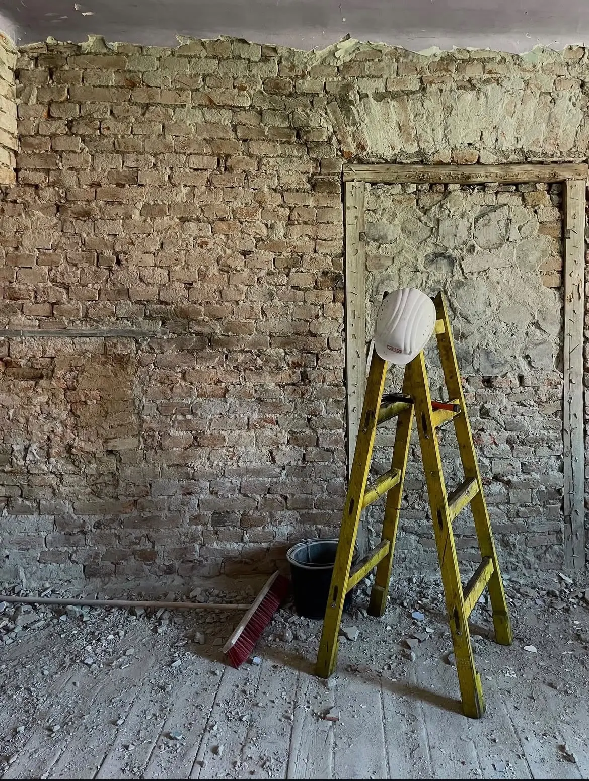 A room stripped back to brick and stone masonry, with a stepladder, helmet, bucket and rubble on the plank floor.